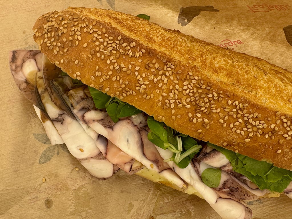close up of sesame seed panino filled with sliced cooked octopus and salad